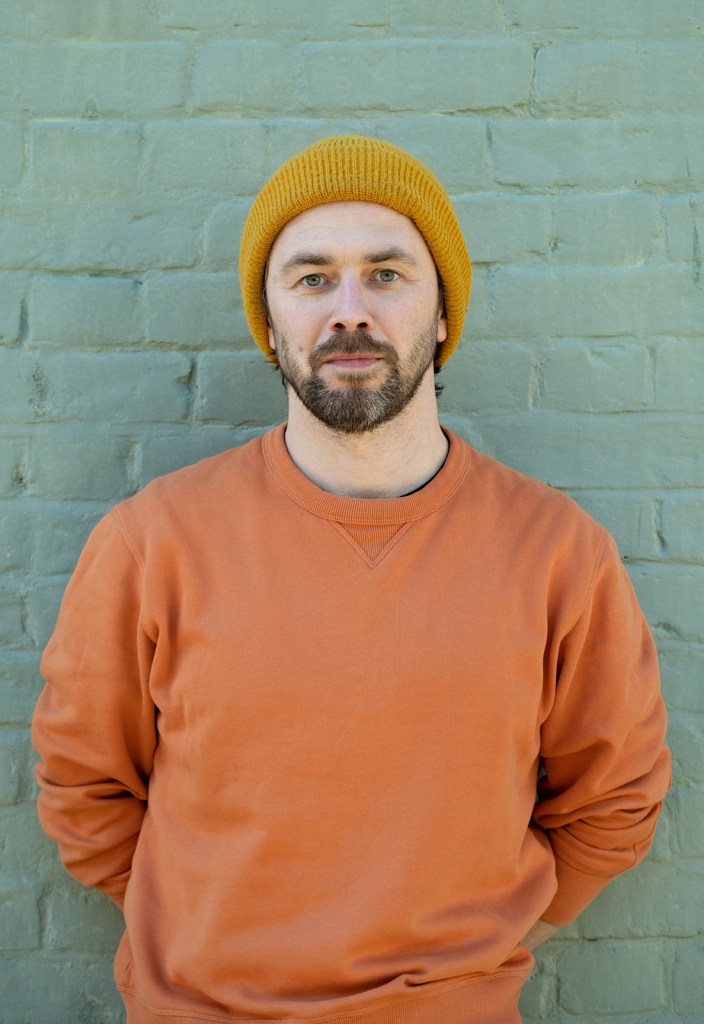 A writer in an orange jumper wears a hat, he looks to the camera and has a brown stubble beard