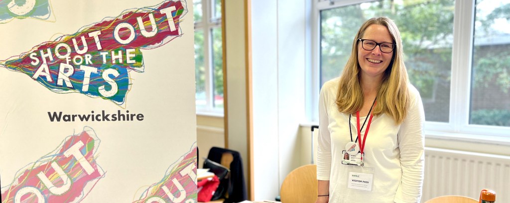 A person with glasses and long blonde hair stands by a table and smiles to the camera