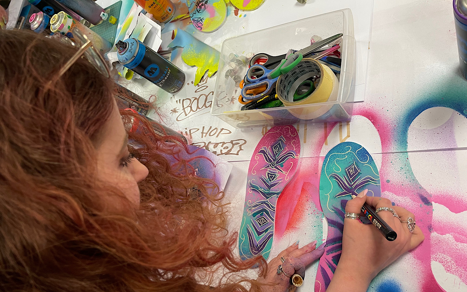 An artist with long red hair is at a table using a gold pen to put patterns on a canvas they have painted pink, blue, green and purple